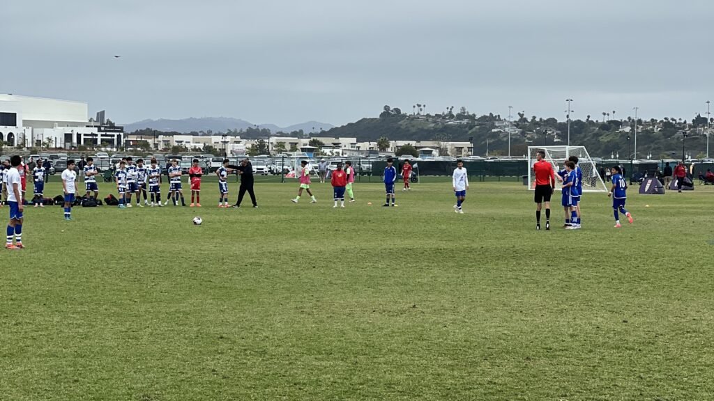 Youth soccer players spread across a grass club field with a coach and a referee